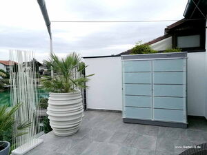 . The image depicts an outdoor area with the following elements:

- A large, white, ribbed planter with a green plant inside.
- A modern, translucent outdoor storage cabinet against a white wall.
- Decorative vertical rods on the left, creating a partition or artistic element.
- A tiled floor.
- Visible neighboring buildings in the background, suggesting a residential setting.