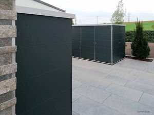 . Zwei moderne, dunkle Gartenhäuser auf einer gepflasterten Terrasse mit Pflanzen im Hintergrund.