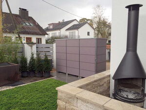 . The image displays a residential outdoor scene with the following elements:

1. **House**: A house with a pitched roof and a skylight.
2. **Outdoor Chimney/Fireplace**: A freestanding metal outdoor chimney or fireplace is visible on the right side.
3. **Garden**: There is a small garden area with neatly trimmed grass.
4. **Potted Plants**: Several potted plants or small trees are placed near a fence.
5. **Storage Unit**: A rectangular storage unit or shed is visible, possibly used for outdoor storage.
6. **Fence**: A modern, horizontal slat fence divides areas of the garden or property.
7. **Other Houses**: Additional houses are visible in the background, suggesting a neighborhood setting.
