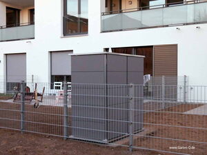 . The image shows a modern, cube-like garden storage unit within a fenced area. It's located in front of a building with a minimalist design. The building has large windows and a balcony with glass railings. The area around the storage unit appears to be a garden or outdoor space with dirt or bare soil.