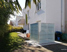 . Hausgarten mit Glasbox, Terrasse, Sitzgelegenheiten und Pflanzen unter blauem Himmel.