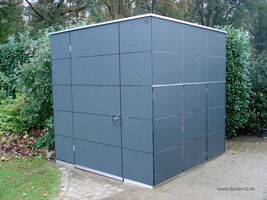 The image shows a modern-looking garden shed with a cube-like structure. It has a sleek, minimalist design with a series of square panels. The shed appears to be located in a garden setting, surrounded by greenery.