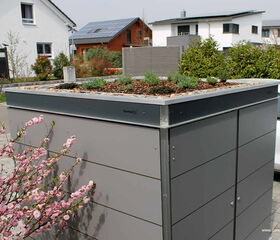 . Ein modernes Gartenmöbel mit bepflanztem Dach, umgeben von Häusern und blühendem Strauch.