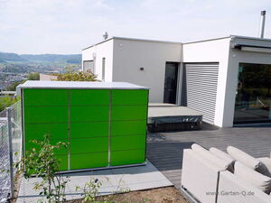 . The image features a modern outdoor space with a green storage unit in the foreground. It is situated on a deck alongside white outdoor furniture. In the background, there is a contemporary building with large windows and a covered hot tub. The setting overlooks a scenic view of hills and a town in the distance.