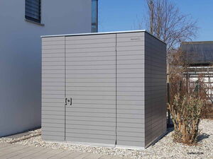 . Ein grauer Gartenschrank steht neben einem Gebäude auf Kiesboden.