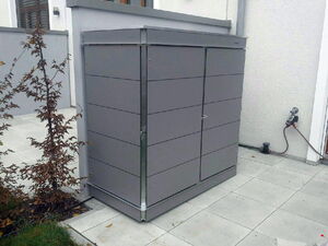 . The image shows a gray metal storage cabinet situated outdoors on a patio area. It's placed near a building wall and is likely used for storing gardening tools, equipment, or other items. There are plants nearby and a water connection with a hose visible on the wall to the right.