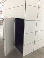 The image shows a large white structure with a grid pattern and an open door, leading to a dark interior. The structure appears to be made of panels and is situated on an indoor concrete floor. The purpose or function of the structure is not clear from the image.