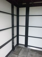 The image shows an interior corner of a structure with a grid pattern of black beams and white panels. The floor is tiled, and the overall look is minimalistic and geometric.