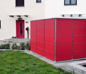 . The image shows a modern residential setting with a red storage unit in the foreground. The storage unit has a sleek, geometric design with metal framing and panels. It's placed on a paved area next to some greenery. In the background, there's a building with white walls, red door, windows with dark frames, and a nearby mailbox. There are also neatly trimmed bushes and a grassy area.