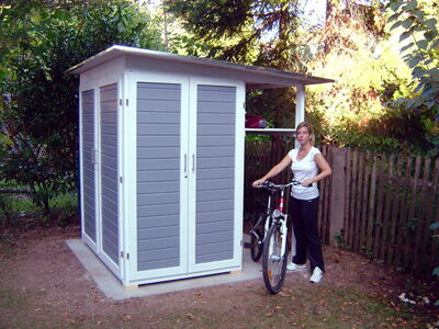 ```json
[
  {
    "object": "shed",
    "description": "A small, modern shed with gray and white paneling, featuring two doors."
  },
  {
    "object": "bicycle",
    "description": "A standard bicycle, being held by a person, likely used for outdoor activity."
  },
  {
    "object": "person",
    "description": "An individual standing next to the bicycle, wearing casual attire."
  },
  {
    "object": "trees",
    "description": "Various trees and foliage surrounding the area, suggesting a garden or backyard."
  },
  {
    "object": "fence",
    "description": "A wooden fence in the background, indicating a boundary of the property."
  }
]
```