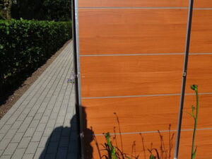 . The image shows part of a structure with wooden panels and a metal frame. There's a paved walkway next to it, bordered by neatly trimmed hedges. In the foreground, plants with long green stems are visible. The setting appears to be outdoors, possibly in a residential or office area.