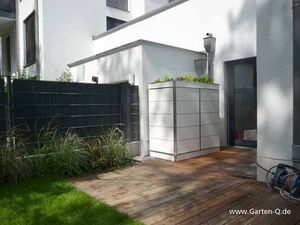 . Terrasse mit Holzboden, weißem Schuppen, schwarzem Zaun, Pflanzen und schlauchartigem Gartenwerkzeug.