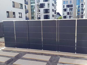 . The image shows a row of solar panels arranged vertically, likely forming a solar fence or barrier. They are positioned in an urban setting with modern residential buildings in the background. The area around the panels has paved pathways and some landscaping.