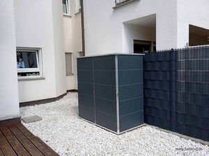 . The image shows an outdoor area with a modern, minimalist storage box placed on white gravel. The storage unit is positioned near a white building wall and a dark fence. The area has a clean, organized appearance.