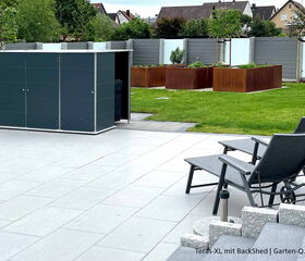 . Ein moderner Gartenschuppen auf einer Terrasse mit Liegestühlen und Hochbeeten im Hintergrund.