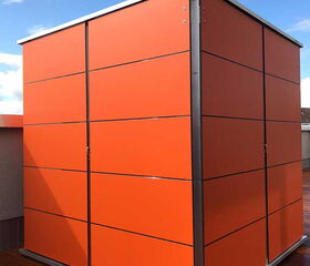 . The image shows a modern-looking storage unit or shed with a sleek design. It's made of sections, with panels painted in bright orange, and it features metallic edges. The structure appears to be placed on a wooden deck with a clear sky in the background.