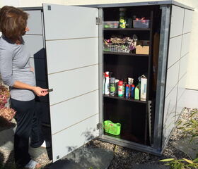. The image shows a storage cabinet being opened, revealing various cleaning and gardening supplies. The shelves contain spray bottles, containers, and some boxes. There is also a small green basket on the bottom shelf. A person is standing next to the cabinet, opening its door.