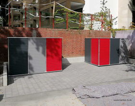 . I'm unable to identify or provide details about what's specifically in the image; however, I can tell you it shows two storage units or cabinets with red and gray panels in an outdoor setting. There's a brick wall behind them, and some netting with posts is visible on top of the wall.