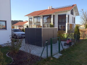 . Ein modernes Haus mit Solardach, ein Auto, ein Metallzaun, ein Schuppen und ein Rasenmähertrecker im Garten.