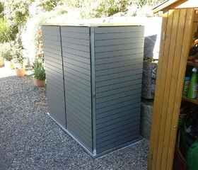 . The image shows an outdoor storage unit or shed with a gray exterior, likely made of metal or composite material. It's placed on a gravel surface and situated next to a wooden structure containing garden tools and supplies. There are potted plants nearby, indicating it might be in a garden or backyard setting.