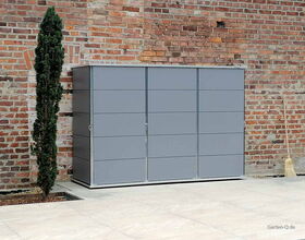 Metallischer Gartenschrank neben einem Baumschössling, rote Ziegelwand im Hintergrund, Besen an der Seite.