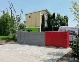 Moderne Gartenboxen in Grau und Rot, gepflasterter Bereich, Gebäude und Bäume im Hintergrund.