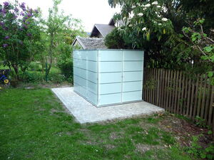 . The image shows a small, modern glass structure with frosted panels, possibly a shed or storage unit, situated in a garden. It is surrounded by grass, trees, flowers, and a wooden fence. There's also a paved area where the structure is placed.