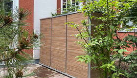 . The image shows a modern garden storage shed with a wood-like finish. It is placed against an exterior wall of a house, surrounded by greenery, including potted plants and a tree. The house has a red and white exterior with windows. The ground is covered with stone pavement.