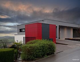 Modernes, flaches Haus mit grauer und roter Fassade, umgeben von grünen Büschen, unter einem dramatischen Himmel.
