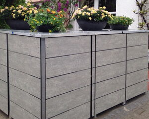 . The image shows a closed storage unit or shed in a garden setting, with a flat top that has flower pots placed on it. The unit is made of materials that resemble gray wood, and it's situated near a house with some plants and flowers around it.