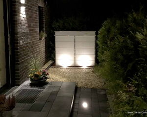 . Ein beleuchteter Gartenweg mit Pflanzgefäß, Ziegelwand und Sträuchern, führt zu einem weißen Schrank im Freien.