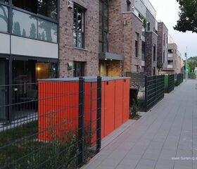 Moderne Gebäude mit Ziegelsteinfassade, Gehweg, Zaun und orangenem Mülltonnenumschlag.