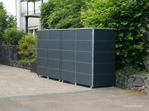 . The image shows a modern, outdoor storage unit or garden shed with a sleek, dark-colored design. It's placed on a paved area next to a building and surrounded by greenery and hedges. The unit has a grid-like panel structure with a simple and clean aesthetic.