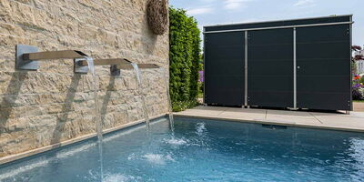 Ein moderner Pool mit Wasserfall an einer Steinmauer und einem schlichten, schwarzen Schuppen daneben.