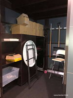 The image shows a storage area with:

1. Cardboard boxes stacked on a shelf.
2. A folding table leaning against the shelf.
3. A folding chair.
4. Long-handled tools like a rake and a broom.
5. Shelving units with various items, including a plastic bin.

The space appears organized for storage purposes.