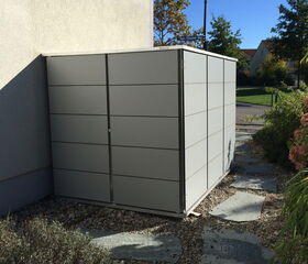 . The image shows a modern, minimalist outdoor storage shed positioned next to a building. The shed appears to be made of metal panels. It is situated on a stone or gravel path, surrounded by some greenery and a small garden. There is a lamp post and more trees in the background. The sky is clear, indicating a sunny day.