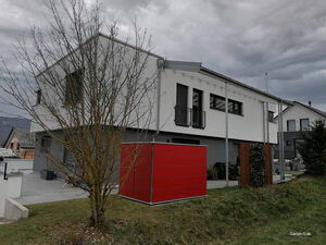 . Modernes Haus mit rotem Gartenhäuschen, trübem Himmel, kahlem Baum und Rasenfläche.