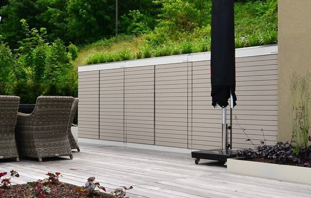 . Gartenmöbel auf Terrasse mit Holzfußboden, dahinter Sichtschutzzaun, schwarzer Sonnenschirm, umgeben von Pflanzen.