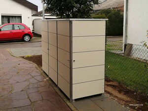 . Ein weißer Geräteschrank steht im Freien neben einer Einfahrt mit einem roten Auto im Hintergrund.
