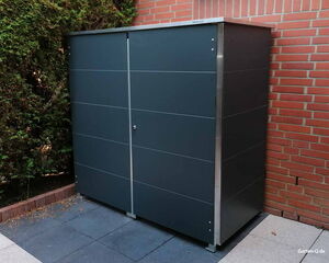 . Ein moderner, dunkelgrauer Gartenschrank steht auf einer Terrasse neben einer Ziegelwand und Hecken.