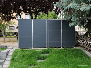 . The image shows a garden with a solid structure, likely a shed or storage unit, that has sleek gray panels. It's surrounded by trees and bordered by a grassy area with stone walkways. The structure is positioned against a backdrop of residential buildings and fencing.