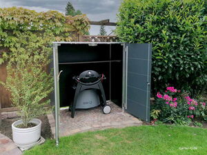 . Ein Weber Grill in einem Gartenschrank, umgeben von Pflanzen und Blumen im Garten.