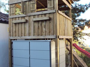 . The image shows a wooden play structure, elevated with a roof, likely a playhouse or treehouse. It has a fenced platform and is supported by pillars. Below the structure is a storage unit with closed cabinets. Nearby, there appears to be the end of a slide, indicating it's a children's play area. Trees and part of a building's roof are visible in the background.