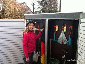 . The image shows a person standing next to an outdoor storage unit containing various garden tools. The tools inside include a rake, a broom, and some other implements neatly organized. The person is wearing a helmet and a pink jacket, smiling at the camera. The storage unit is located outside, with some fencing and trees visible in the background.