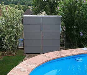 . The image shows a grey storage cabinet situated outdoors, surrounded by a garden with green trees and shrubs. In the foreground, there's a blue swimming pool with a stone or tiled edge. The setting seems to be a backyard or garden area.