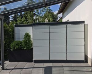 The image shows a modern outdoor setting with a privacy screen or panel system on a patio. The panels have a sleek, grid-like design. There is also a planter with greenery to the left and a portion of a glass or transparent roof above. The setup appears to be against the wall of a house.