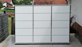 . This image shows a large, modern outdoor storage unit with a grid-like design. It appears to be placed on a patio or paved area beside a house. The background includes some residential houses and greenery.