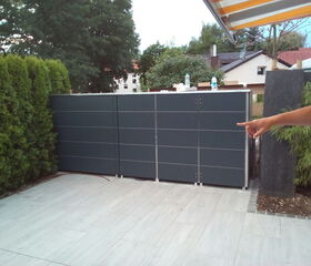 The image shows a backyard or garden area with a section of fencing or a partition that appears to be made of dark, horizontal panels. There's foliage on the left and right sides, and some houses are visible in the background. A person's arm is extended, pointing towards the fence.