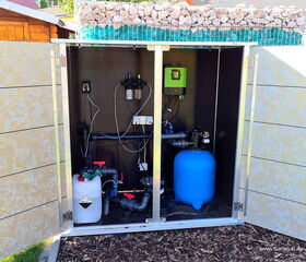 The image shows a setup in an outdoor cabinet containing pool or garden equipment. Here's a breakdown of the components seen inside:

1. **Storage Cabinet**: An open cabinet made of metal or another durable material.
2. **Pumps and Filters**: 
   - A blue cylindrical container, likely a water filter or pump.
   - Pipes and valves for water flow control.
3. **Control Box/Panel**: 
   - A green-colored control unit mounted inside.
   - Several wires and cables indicating electronic control systems.
4. **Chemical Container**: 
   - A white container, possibly for storing pool chemicals or other solutions.
5. **Rock and Glass Cover**: 
   - Rock and glass material on top for decorative or functional purposes.

The setup appears organized and neatly installed for outdoor water management tasks.