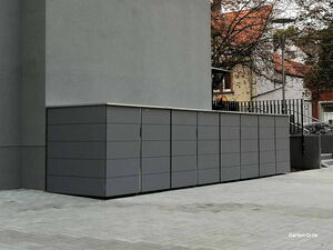 . Graue, moderne Mülltonnenboxen stehen an einer Hauswand in einer städtischen Umgebung.